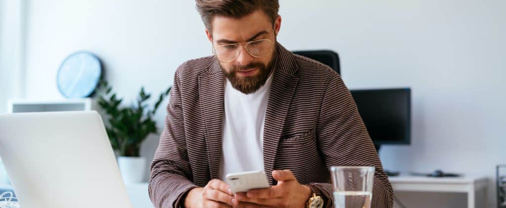 Young Businessman Checking Phone While Working With Laptop In Home Office By Marketing Refresh In Houston, Texas. 